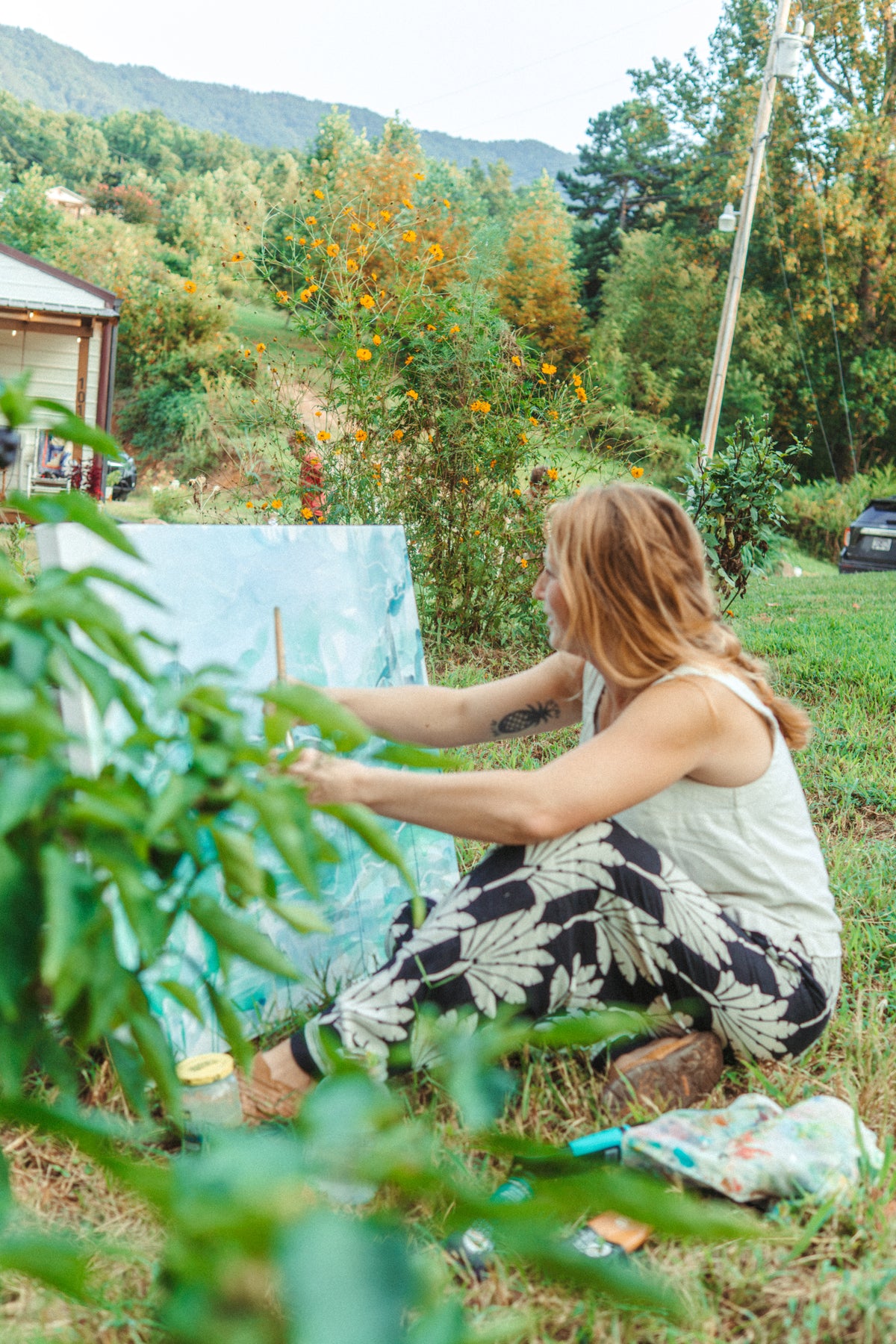 A woman sits on the grass outdoors, painting on a canvas easel. She is wearing a white tank top and black-and-white patterned pants.
